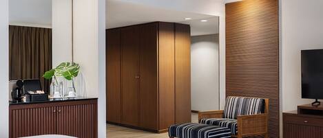 A modern hotel room with a large bed, a wooden wardrobe, a striped armchair, a television, and a coffee machine on the desk.
