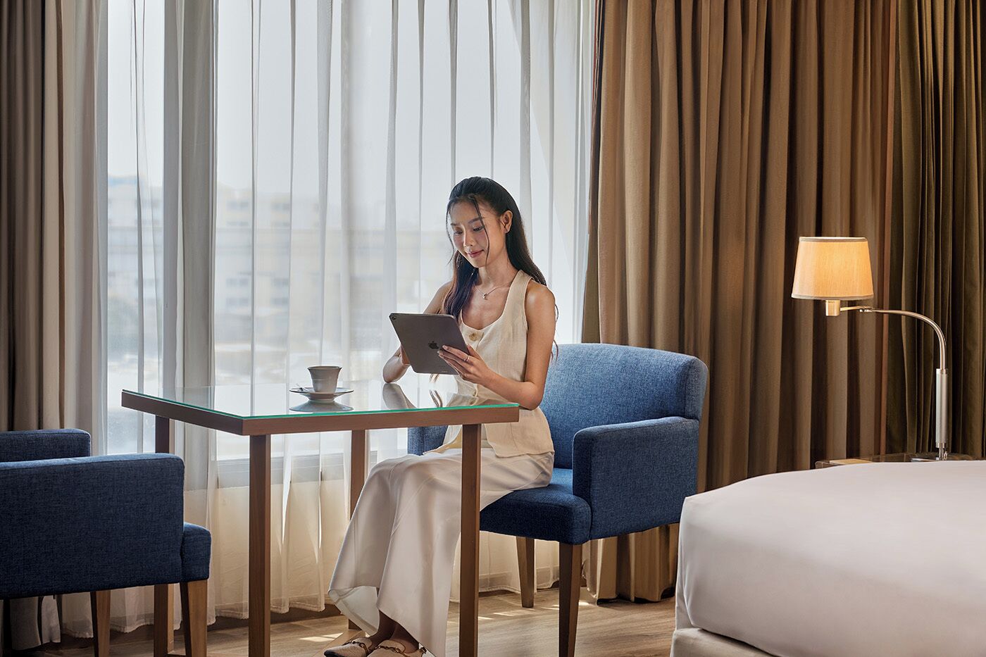 A hotel room with a bed, a blue armchair, a glass table with a cup, and a lamp.