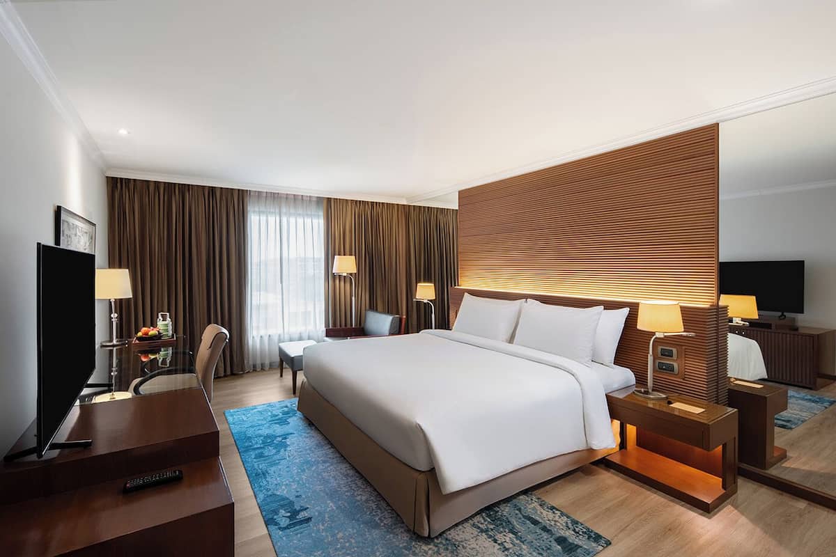 A modern hotel room with a large bed, a desk with a TV, and a seating area with a chair and lamp.