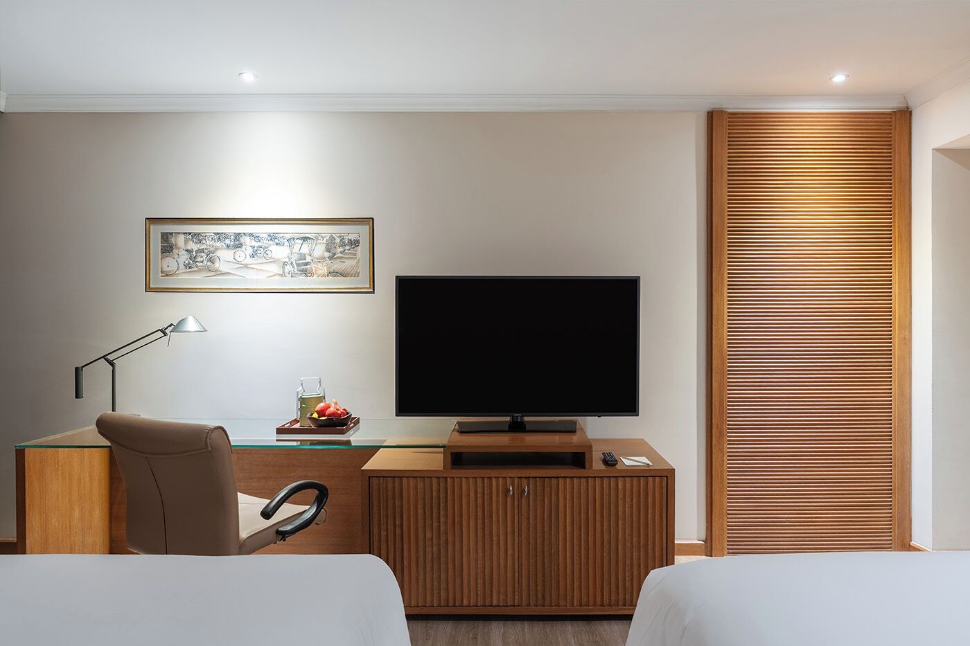 A modern hotel room with a wooden desk, a flat-screen TV, a framed picture, a lamp, and a wooden partition.
