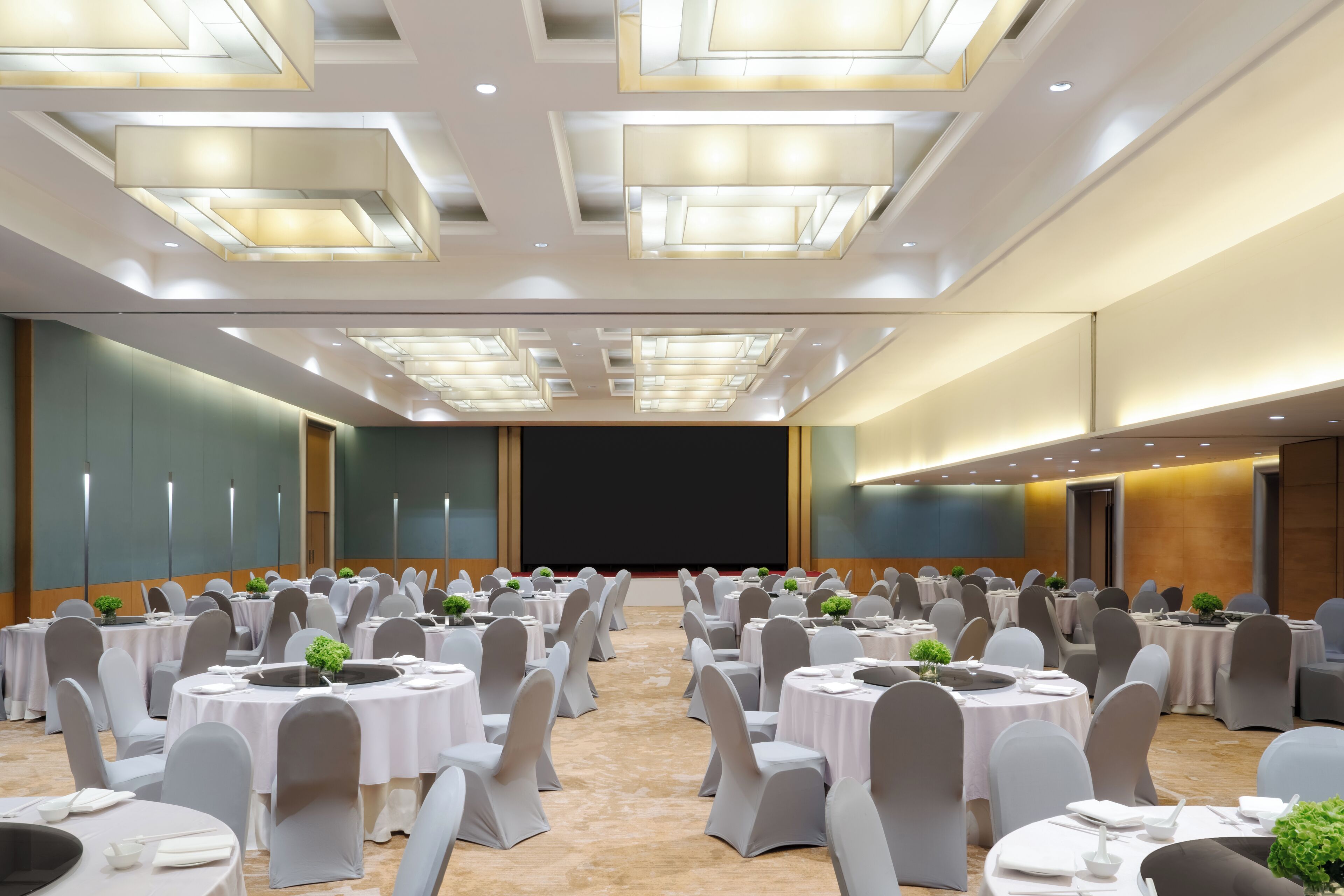 Un salón de conferencias amplio con varias mesas redondas, sillas grises y un escenario con una pantalla grande.