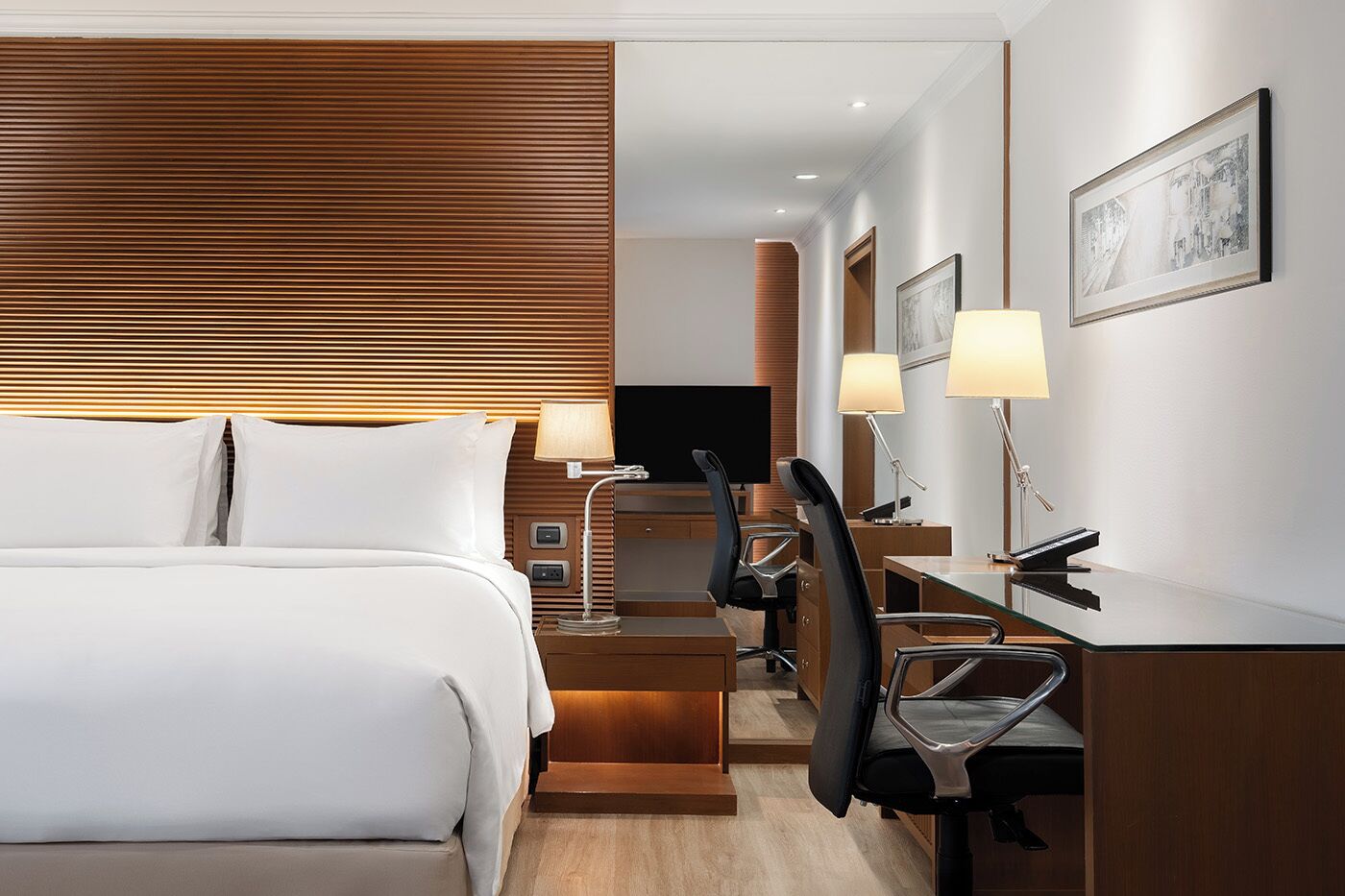 A modern hotel room with a large bed, a desk with two chairs, a television, and a desk lamp.