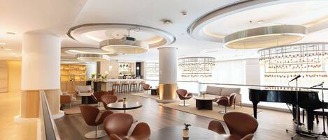 A modern lounge area with round tables, comfortable seating, a grand piano, and a bar in the background.