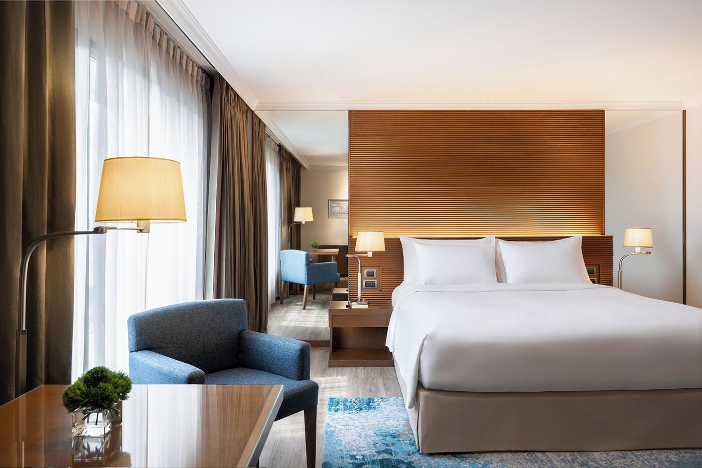 Una habitación de hotel con una cama grande, un escritorio con silla, una mesita con un florero y una silla azul.