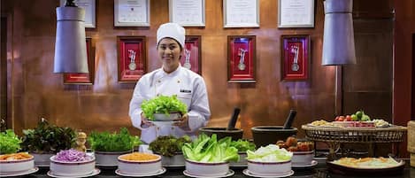 A chef holding a bowl of lettuce in a kitchen with various dishes displayed on the counter.