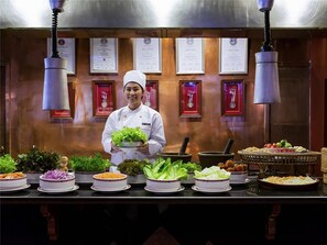 Ein Koch hält eine Schüssel mit Salat in einer Küche, in der verschiedene Gerichte auf der Arbeitsfläche präsentiert werden.