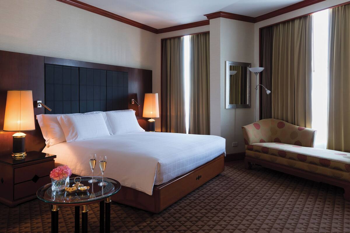 Ein Hotelzimmer mit einem großen Bett, zwei Nachttischlampen, einem kleinen Tisch mit einer Blumenvase und zwei Champagnergläsern, einem Sofa und einem großen Fenster mit Vorhängen.