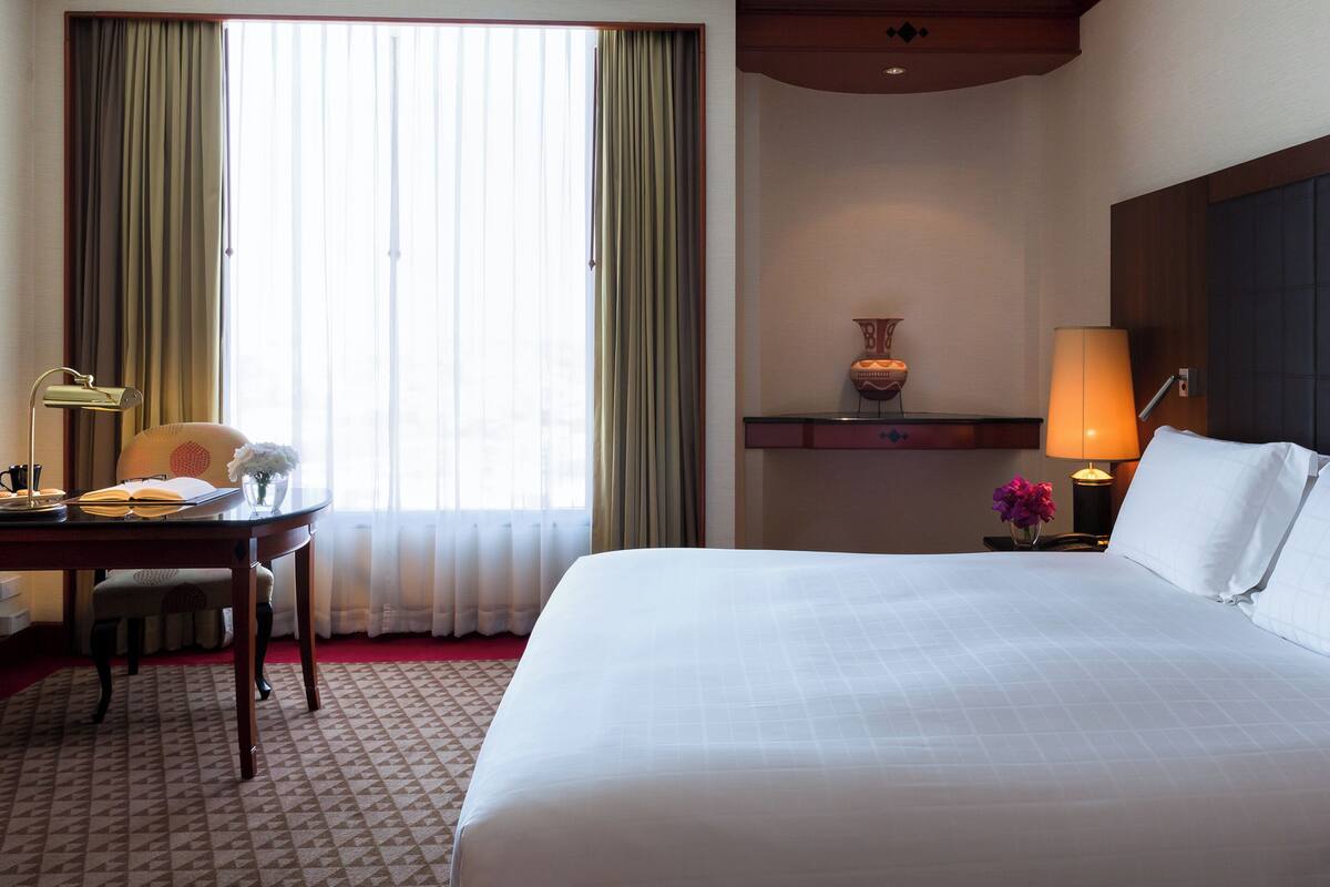 Ein Hotelzimmer mit einem großen Bett, einem Schreibtisch, einem Stuhl, einer Lampe und einer Blumenvase.