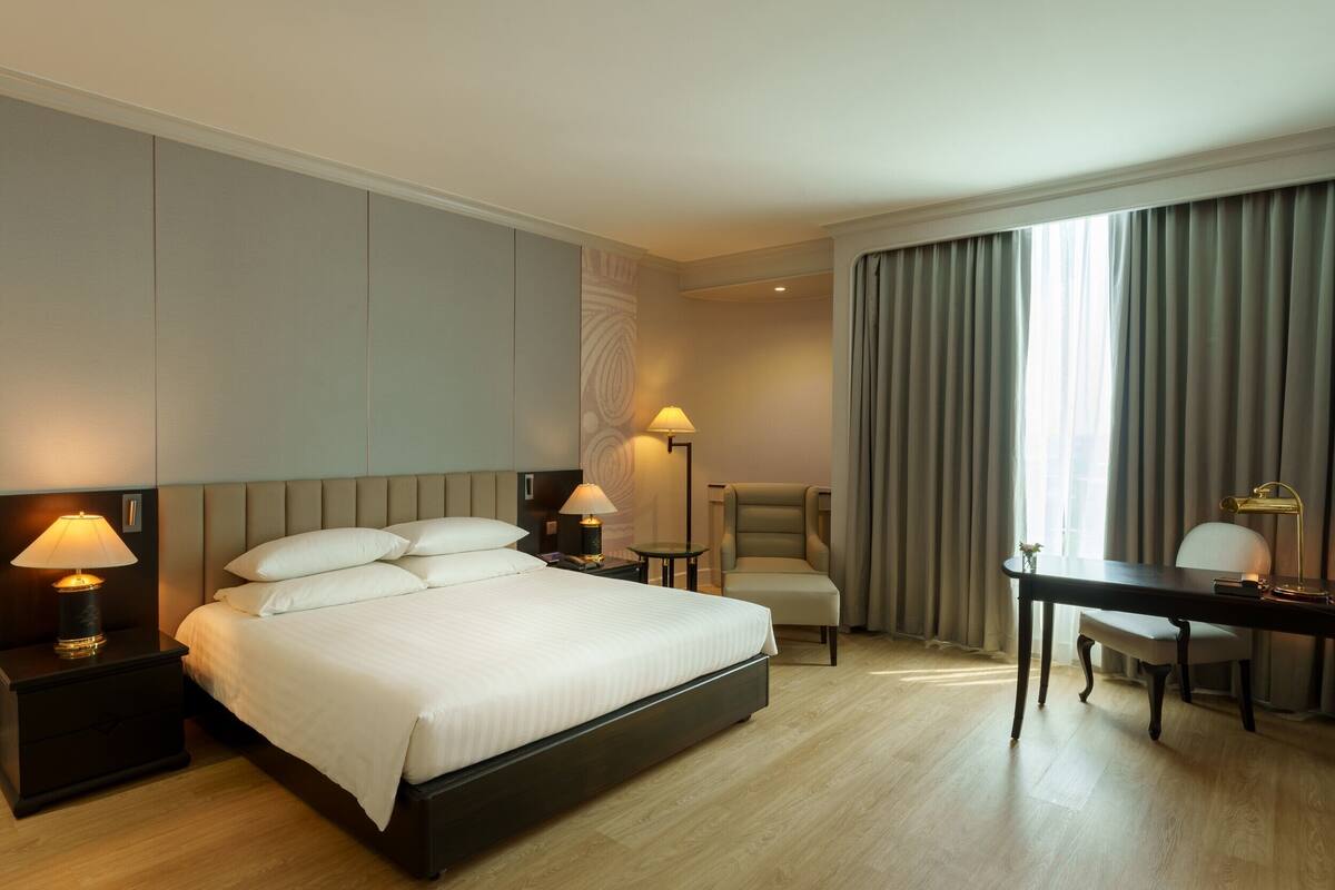 Ein Hotelzimmer mit einem großen Bett, Nachttischen mit Lampen, einem Schreibtisch, einem Sessel und Blick ins Freie durch Vorhänge.