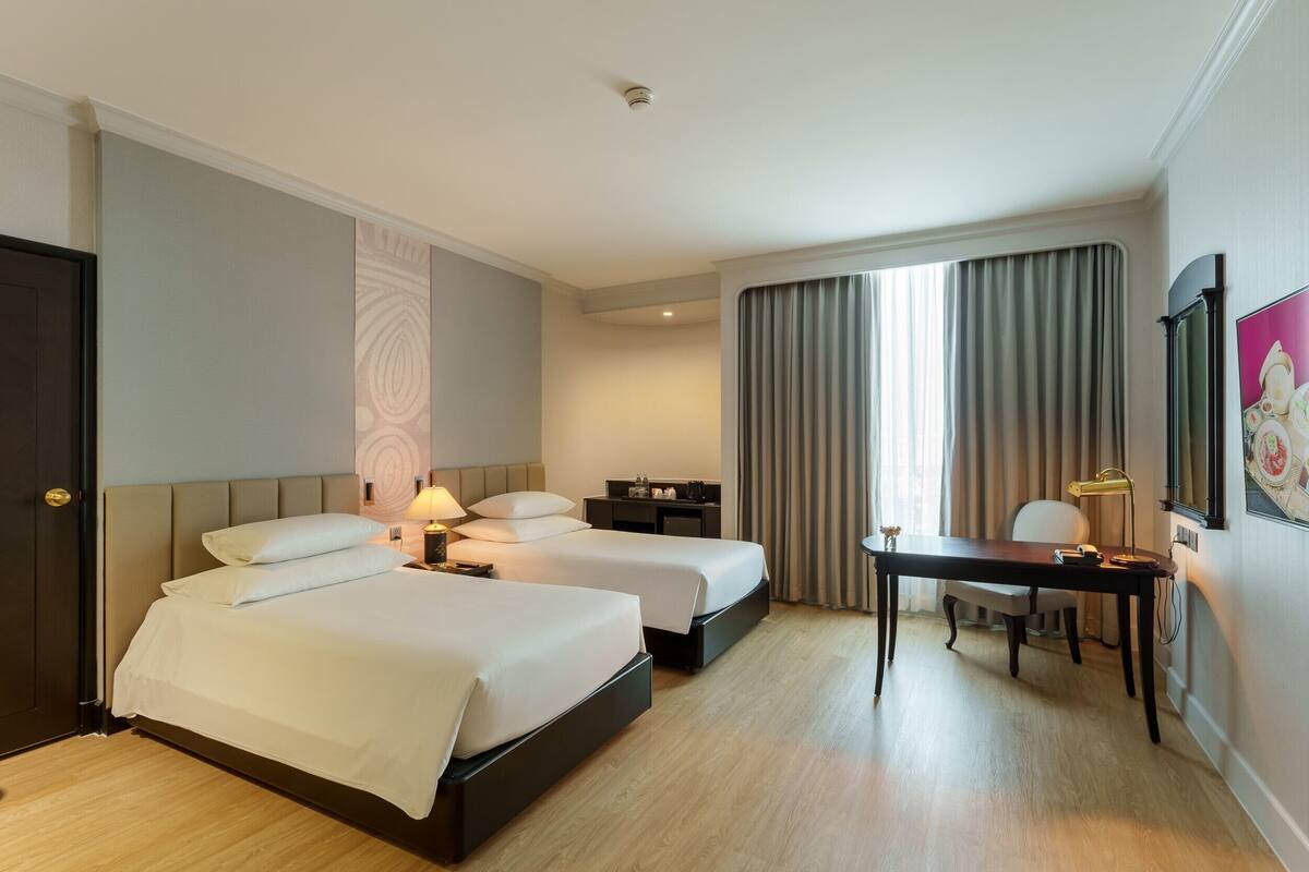 Ein Hotelzimmer mit zwei Betten, einem Schreibtisch, einem Stuhl, einer Schreibtischlampe, einem Nachttisch und einem großen Fenster mit Vorhängen.