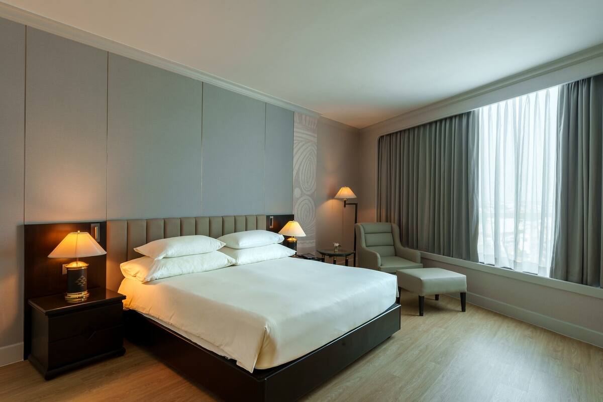 Ein Hotelzimmer mit einem großen Bett, zwei Nachttischlampen, einem Sessel und einem Fenster mit Vorhängen.