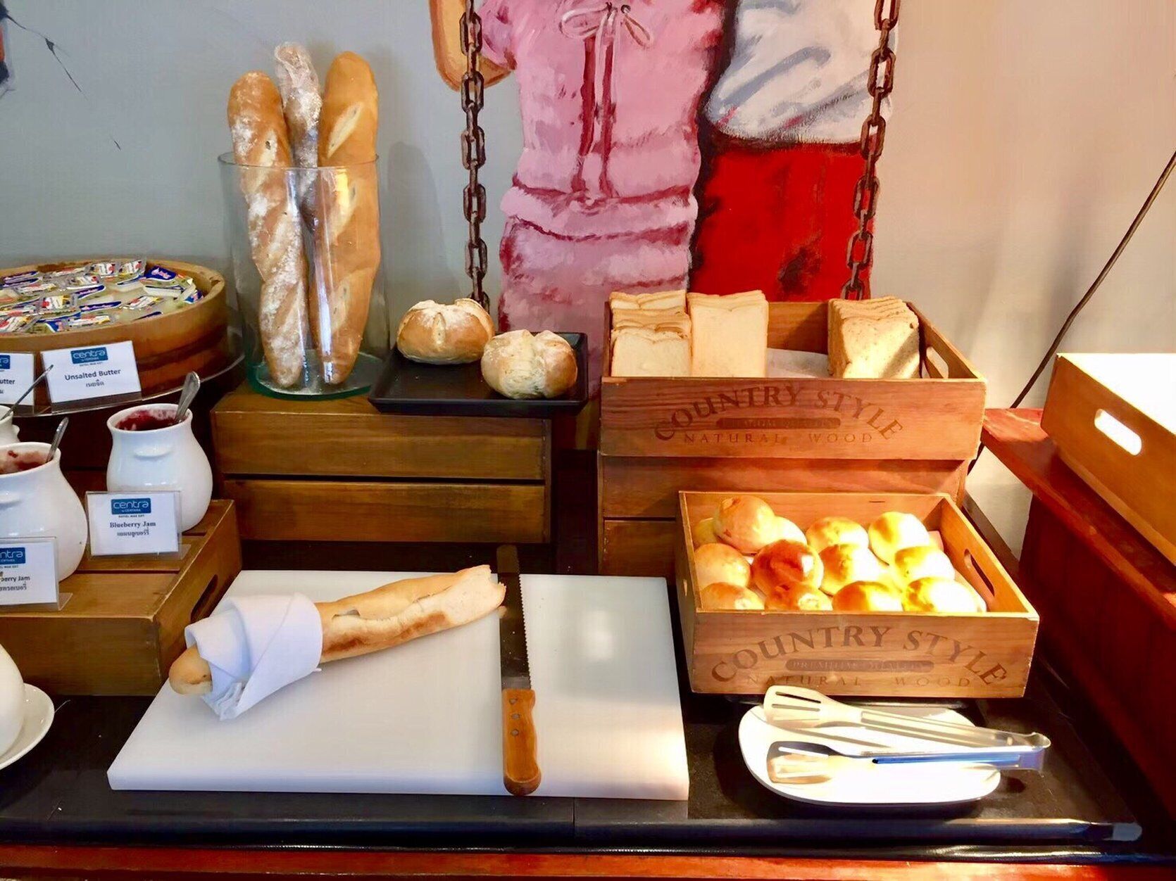 A bakery display with various breads and pastries, a cutting board with a knife, and jars of spreads.