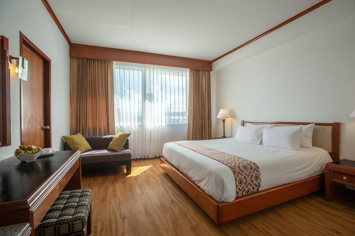 Une chambre d’hôtel avec un grand lit, un bureau avec une chaise, une petite table avec un panier de fruits et une fenêtre avec des rideaux.
