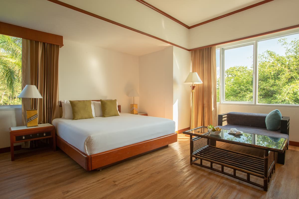 Une chambre d’hôtel avec un grand lit, un canapé, une table basse en verre et une vue sur la verdure.