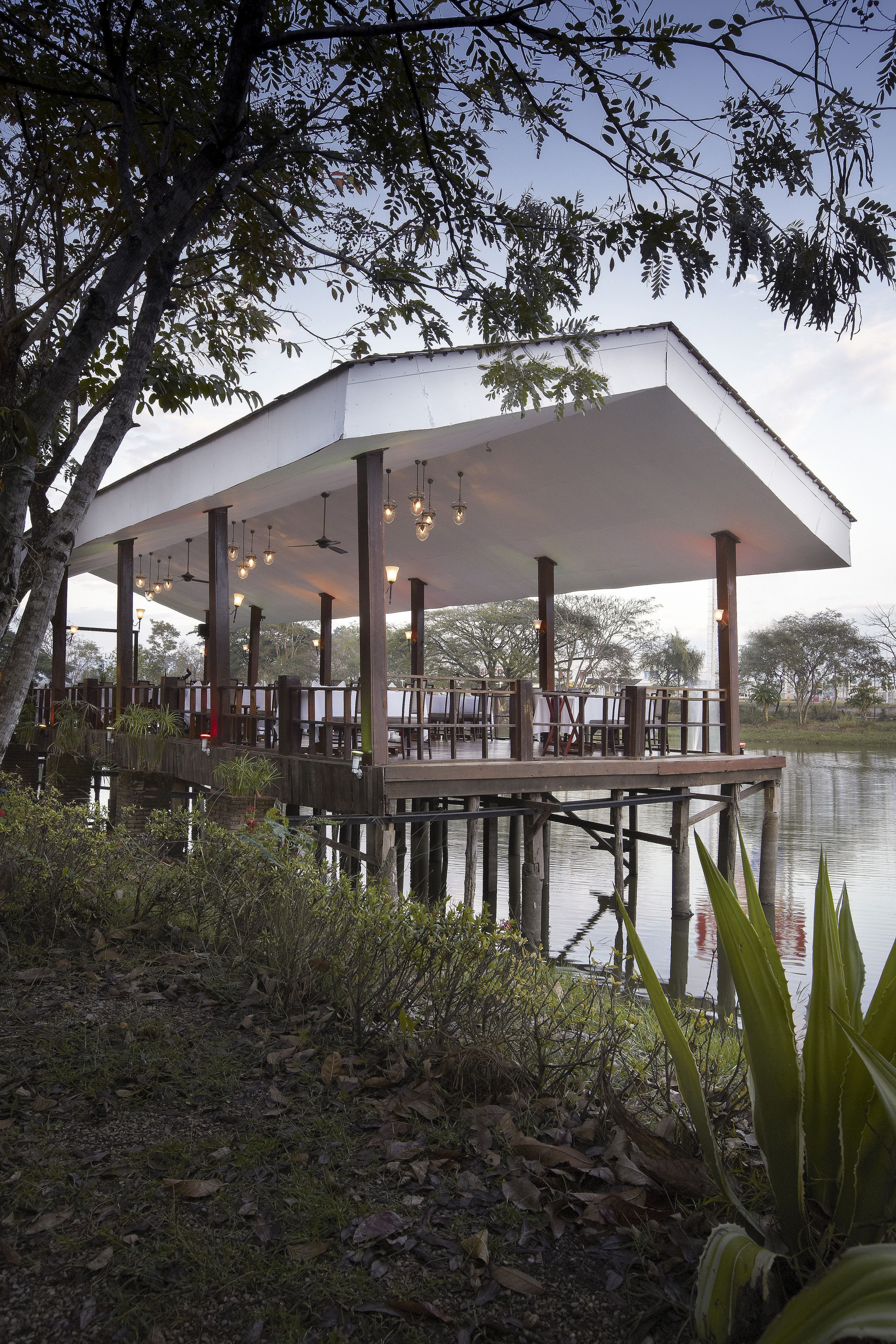 A covered outdoor dining area with wooden pillars and hanging lights, overlooking a calm body of water.