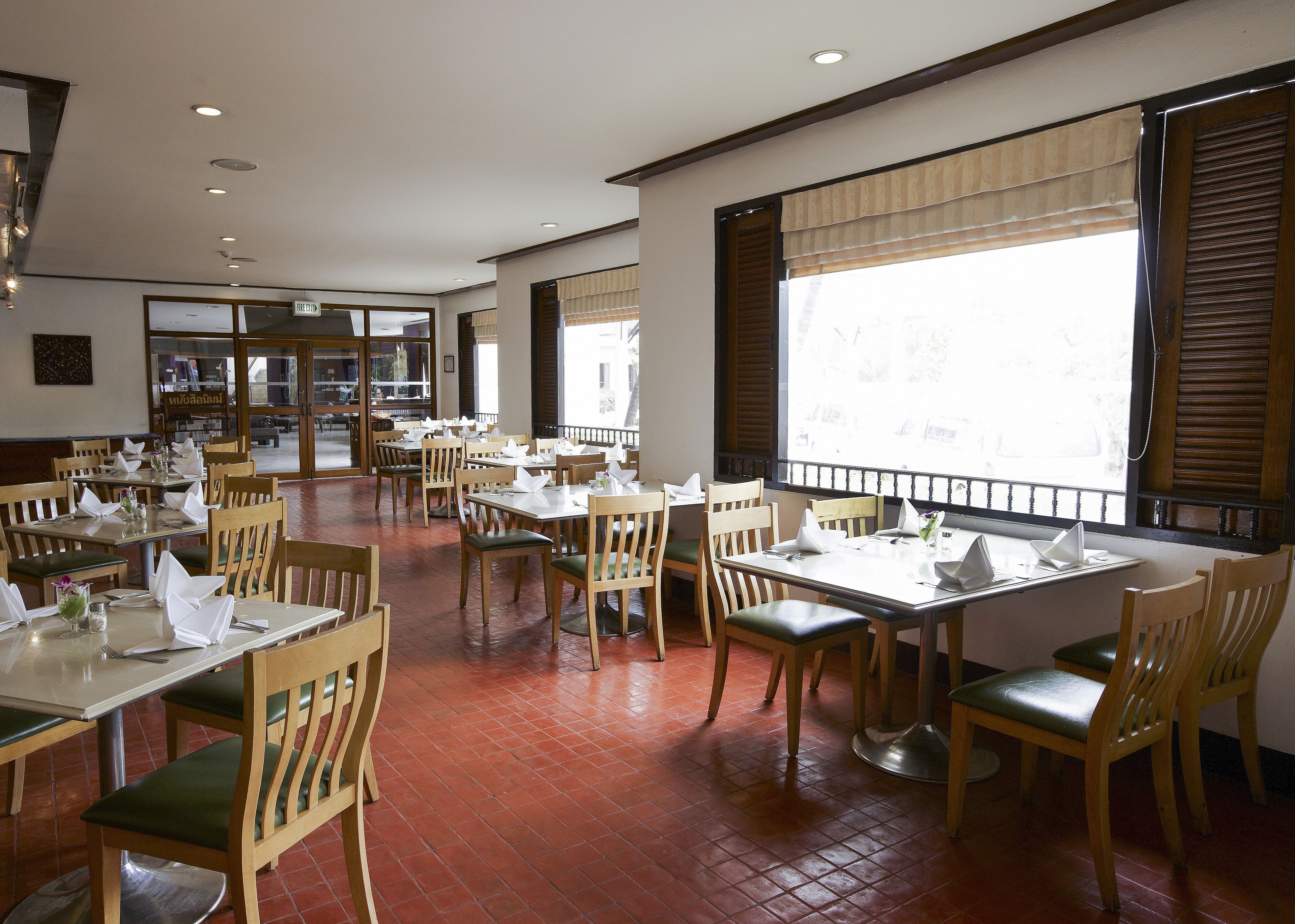 Un restaurant spacieux avec des tables et des chaises en bois, de grandes fenêtres équipées de stores et un sol carrelé.