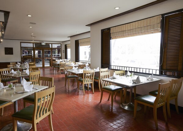 A spacious restaurant with wooden tables and chairs, large windows with blinds, and a tiled floor. - Mae Sot Hotel (Mae Sot)