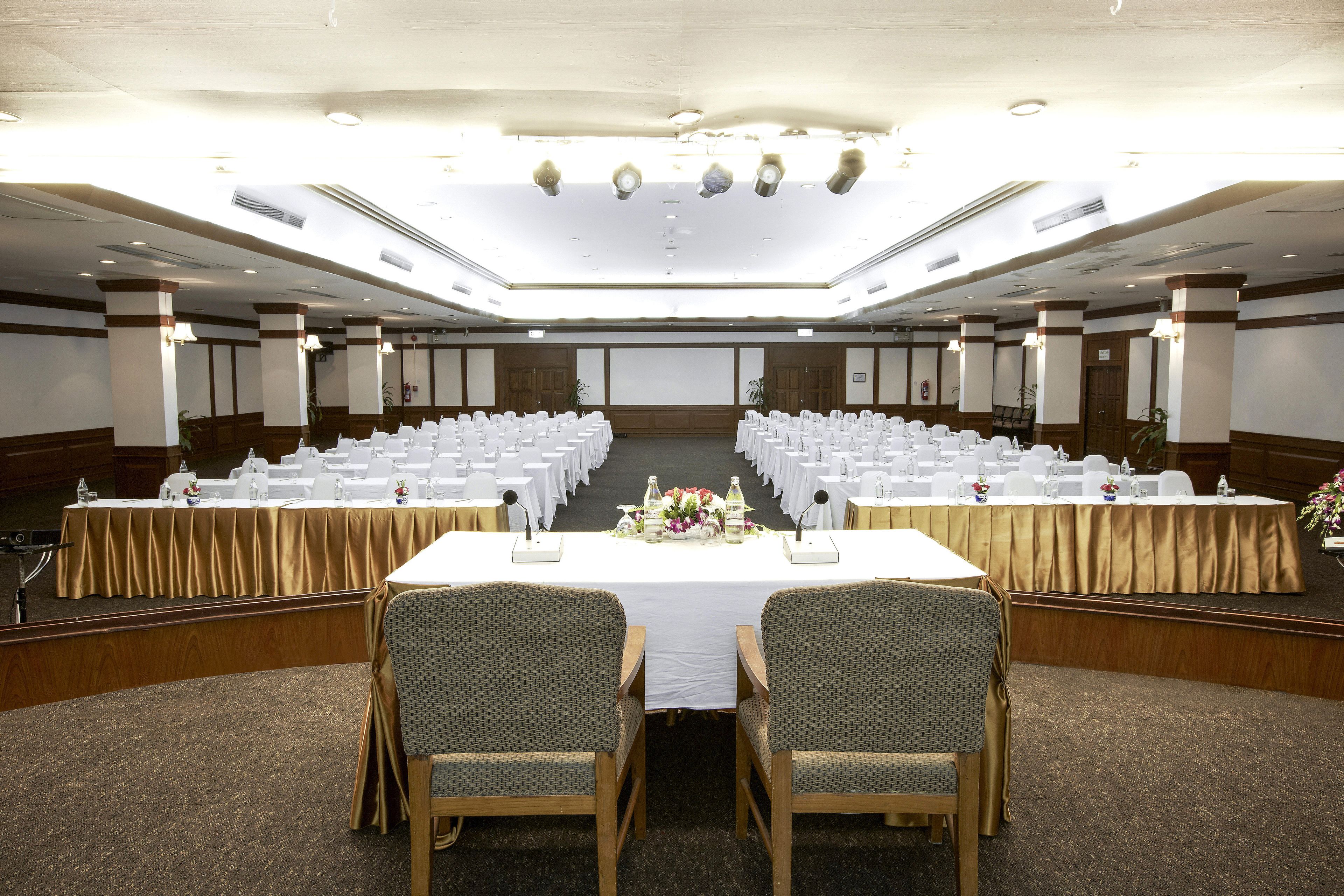 Un amplio salón de conferencias con filas de mesas y sillas blancas, un escenario con un atril y un techo alto con iluminación empotrada.