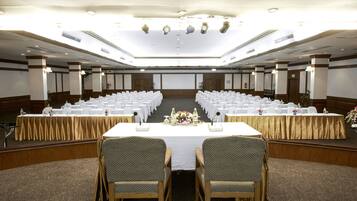 Un amplio salón de conferencias con filas de mesas y sillas blancas, un escenario con un atril y un techo alto con iluminación empotrada.