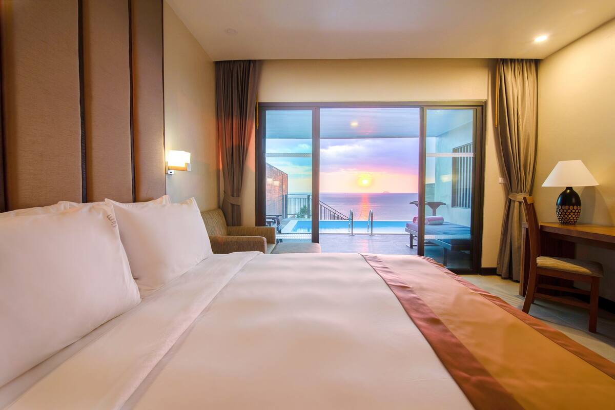 Une chambre d’hôtel avec un grand lit, un bureau, une chaise et une vue sur le coucher de soleil, visible à travers les portes du balcon.