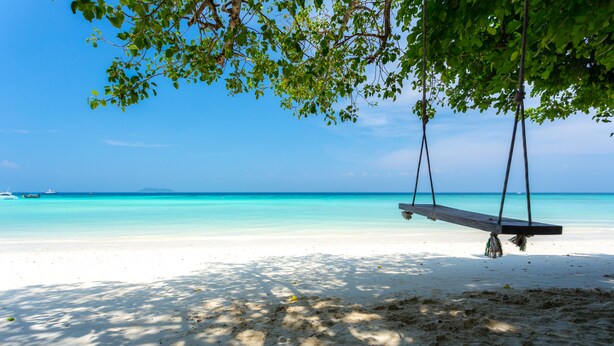 Una playa con una hamaca bajo un árbol, agua turquesa cristalina y un barco a lo lejos.