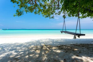 Une plage avec une balançoire sous un arbre, une eau turquoise et limpide, et un bateau à l’horizon.