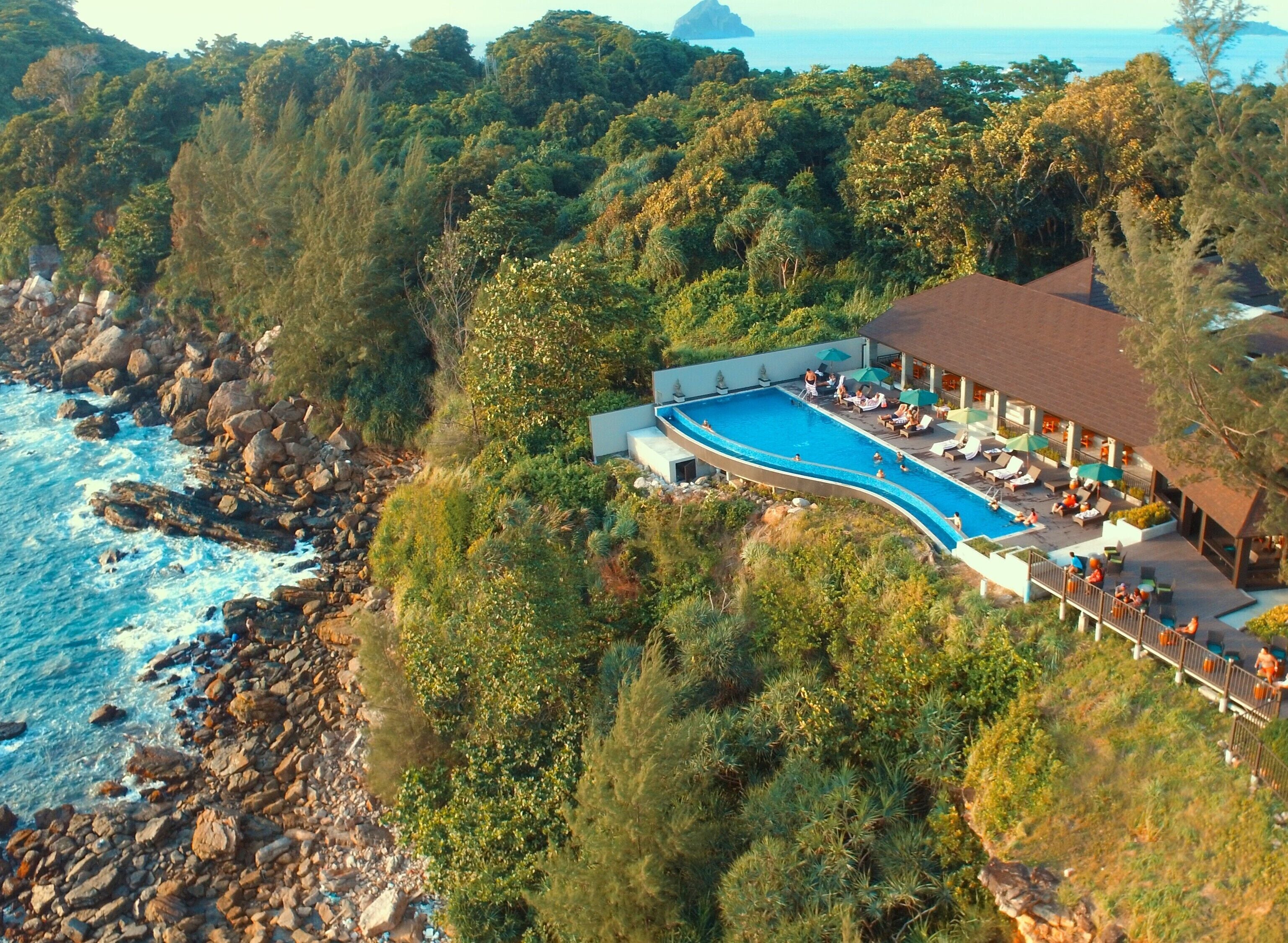 An aerial view of a resort with a swimming pool, surrounded by dense vegetation and rocky coastline.