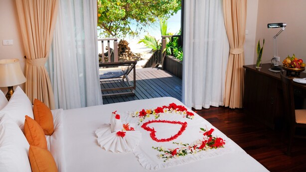 A hotel room with a bed, curtains, a desk with a lamp, and a view of a deck and outdoor area. - Phi Phi Holiday Resort (Ko Phi Phi)