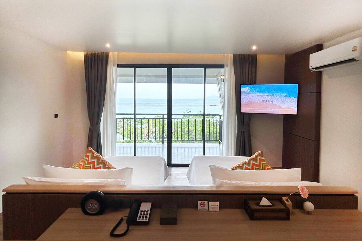 Une chambre d’hôtel avec un lit, un téléviseur fixé au mur, un balcon avec vue sur la mer et un bureau équipé d’un téléphone et d’une horloge.