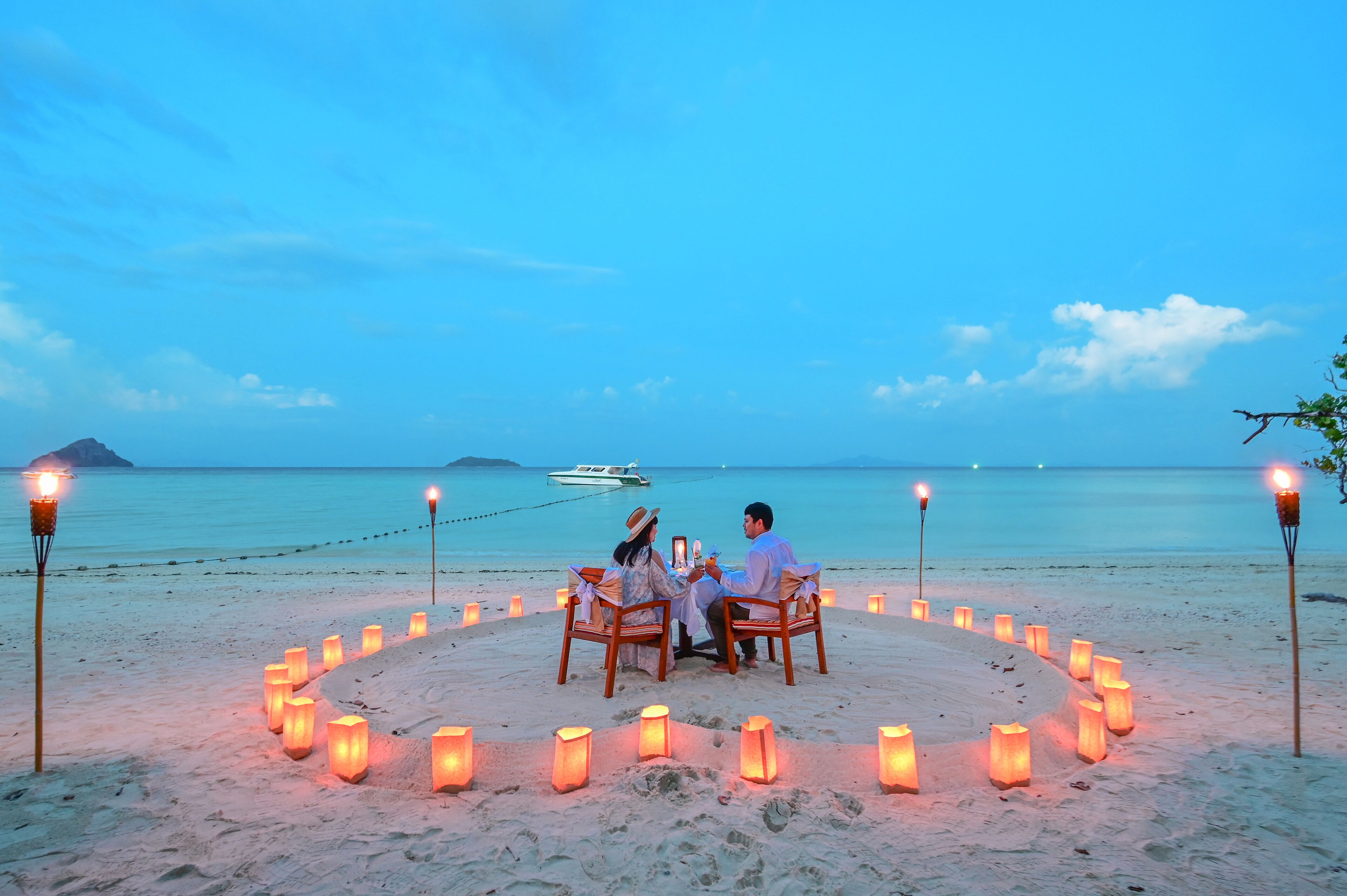 A couple dining at a beachside table with candles, surrounded by a circle of candles, with a boat in the background.