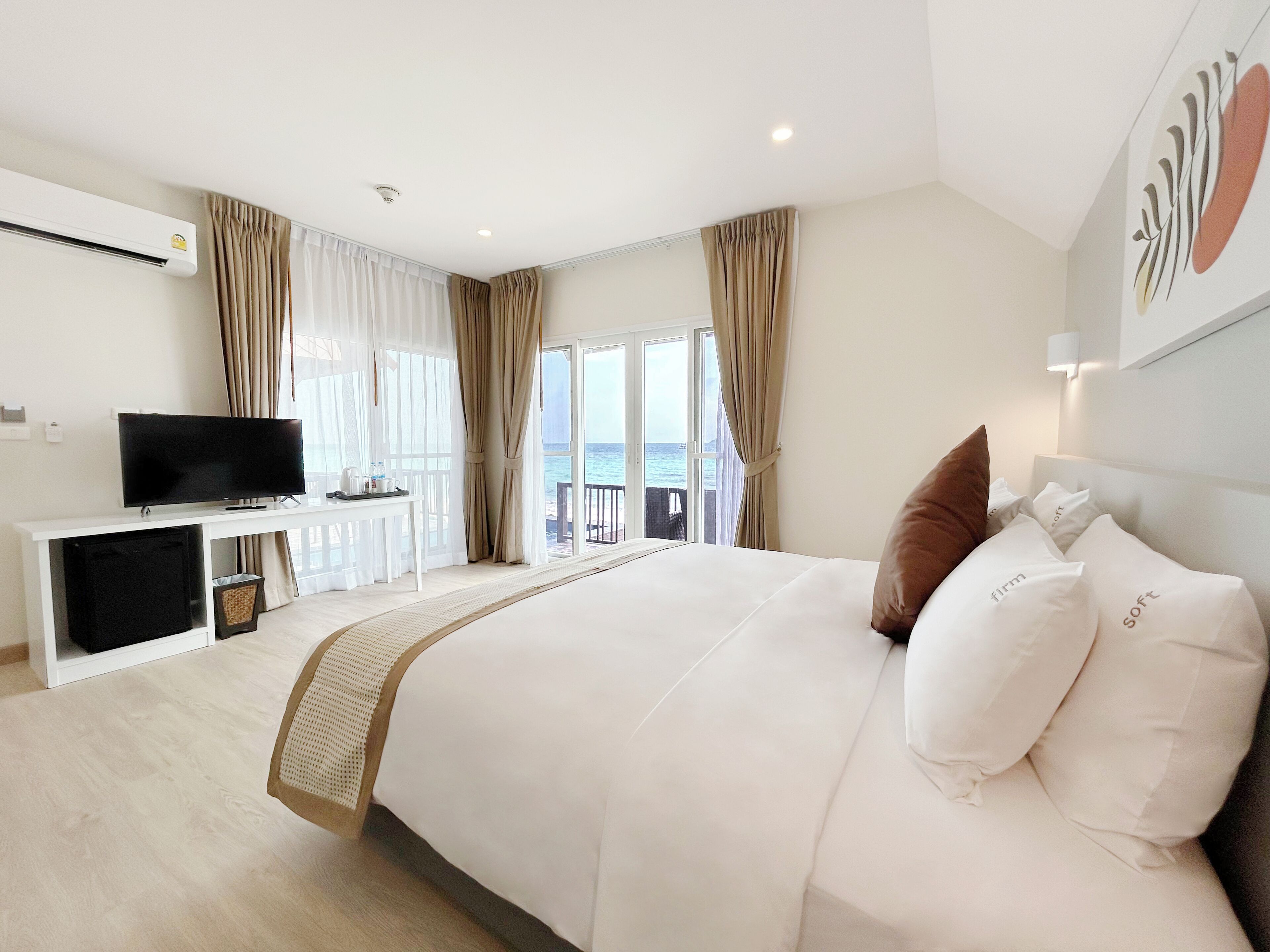 A hotel room with a large bed, a TV on a white console, a desk with a tray of items, and a view of the sea through the windows.