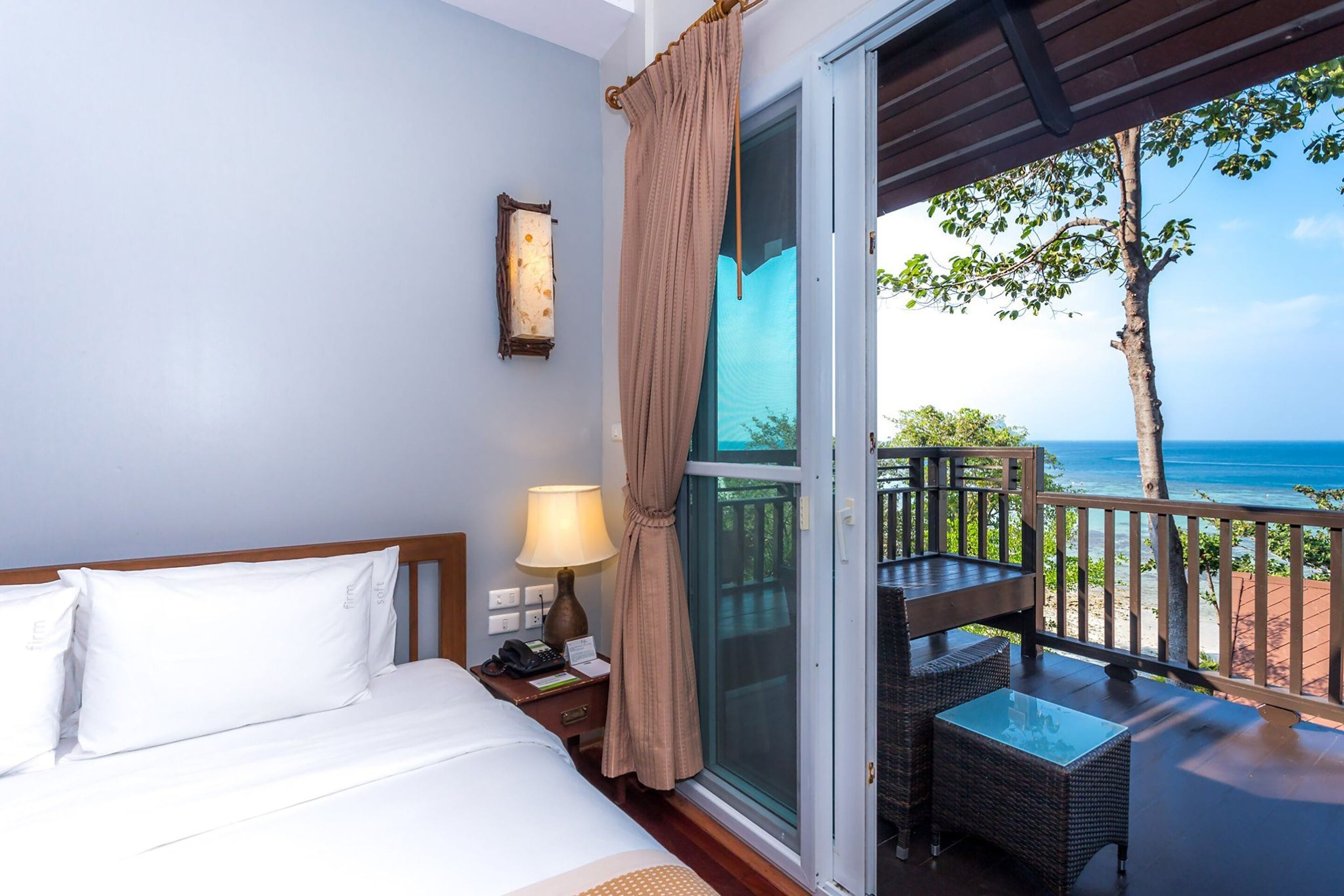 A hotel room with a bed, a nightstand, a lamp, a window with curtains, and a balcony with a table and chairs overlooking the ocean.