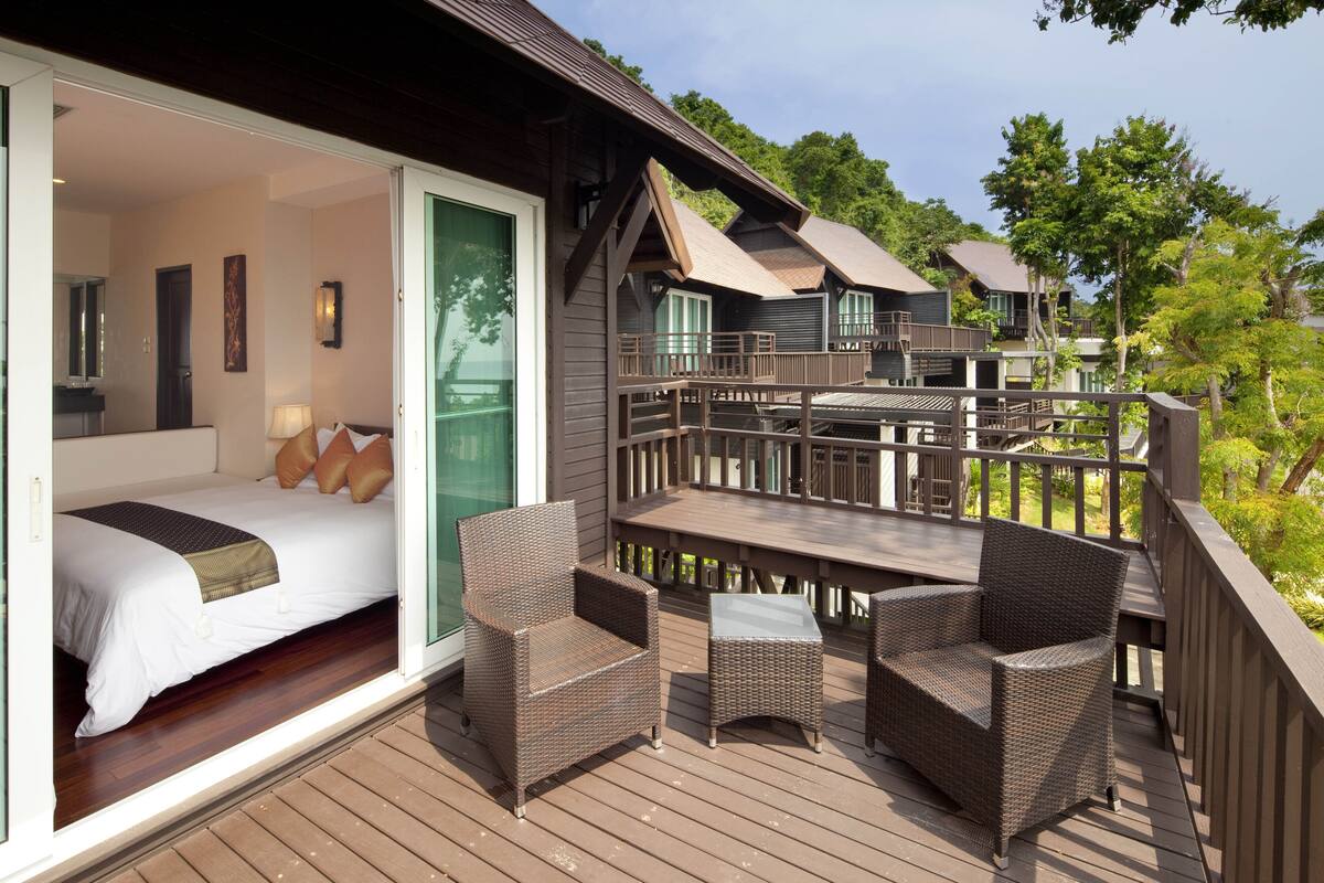 Une terrasse en bois avec des meubles en osier, un lit avec une literie blanche et une porte verte donnant sur une pièce.