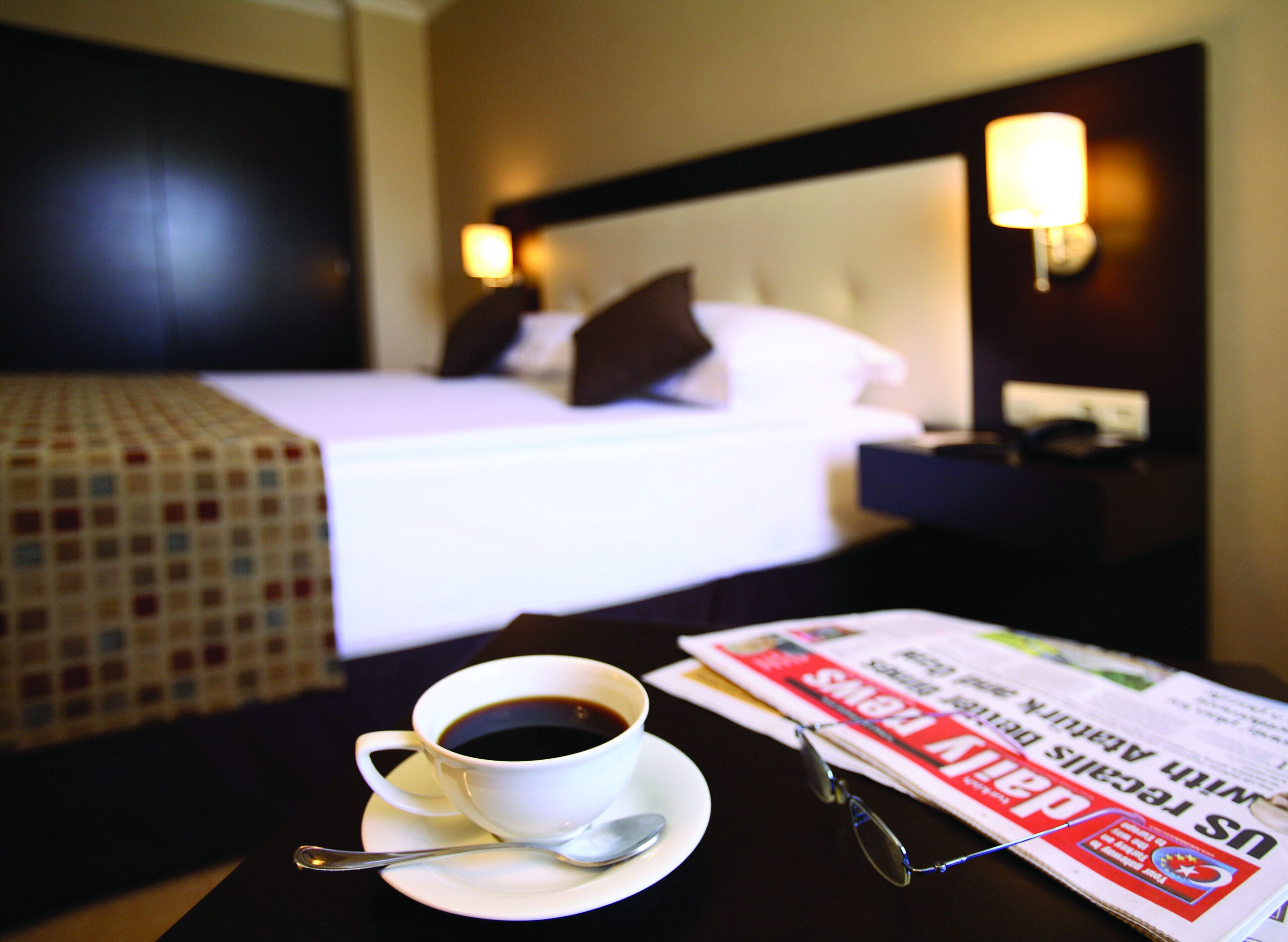 Habitació d'hotel amb llit, làmpades de nit, una tassa de cafè sobre una taula i un diari.