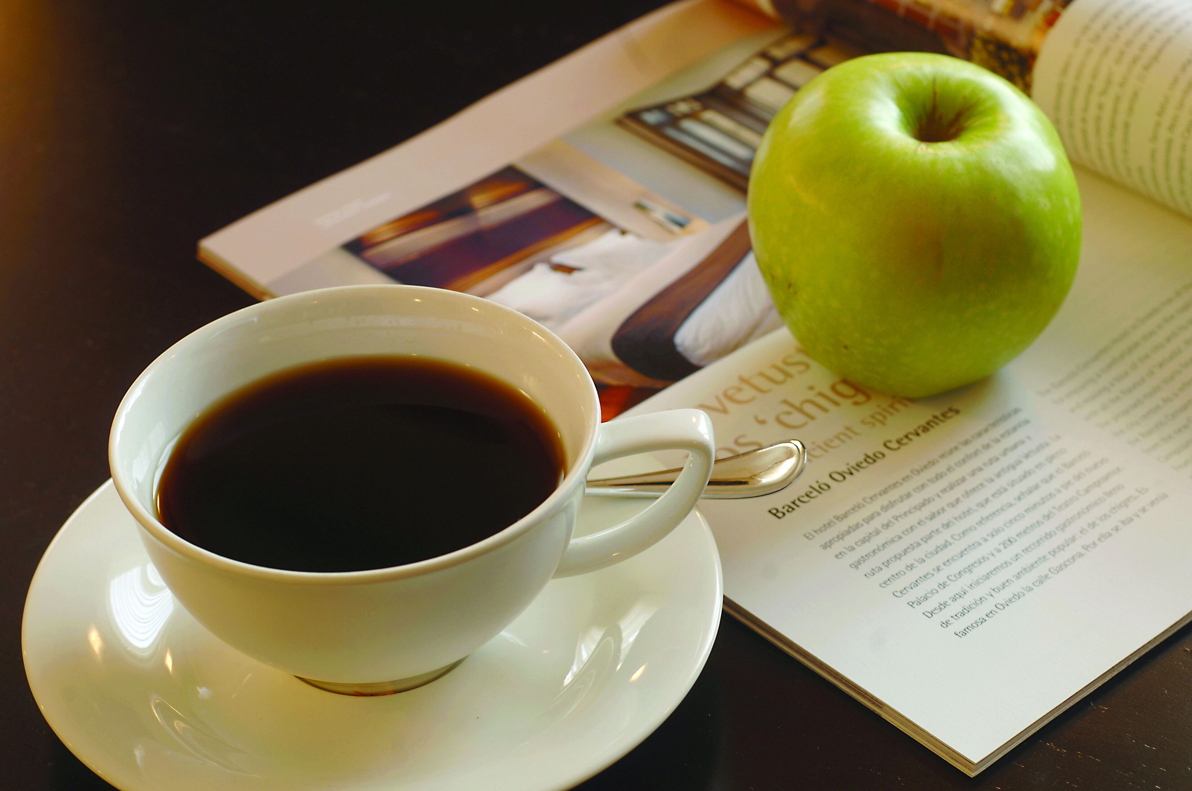 Una taza de café sobre un plato, una manzana verde y una revista abierta sobre una mesa.