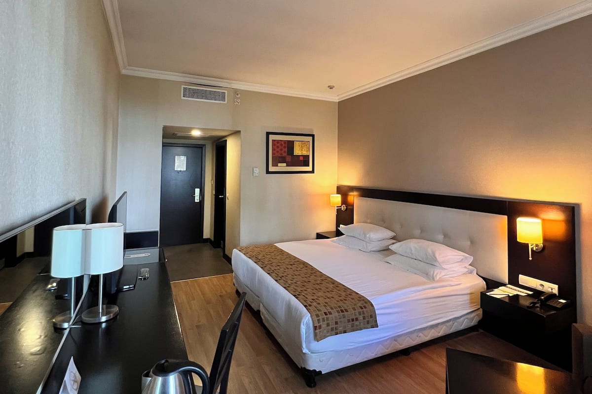 A hotel room with a bed, bedside tables, a desk with a lamp, a chair, a coffee maker, and a painting on the wall.