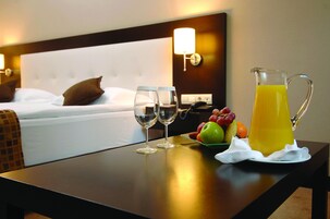 Una habitación de hotel con una cama bien hecha, una mesita de noche con dos vasos de agua, una jarra de jugo de naranja y un plato de fruta.