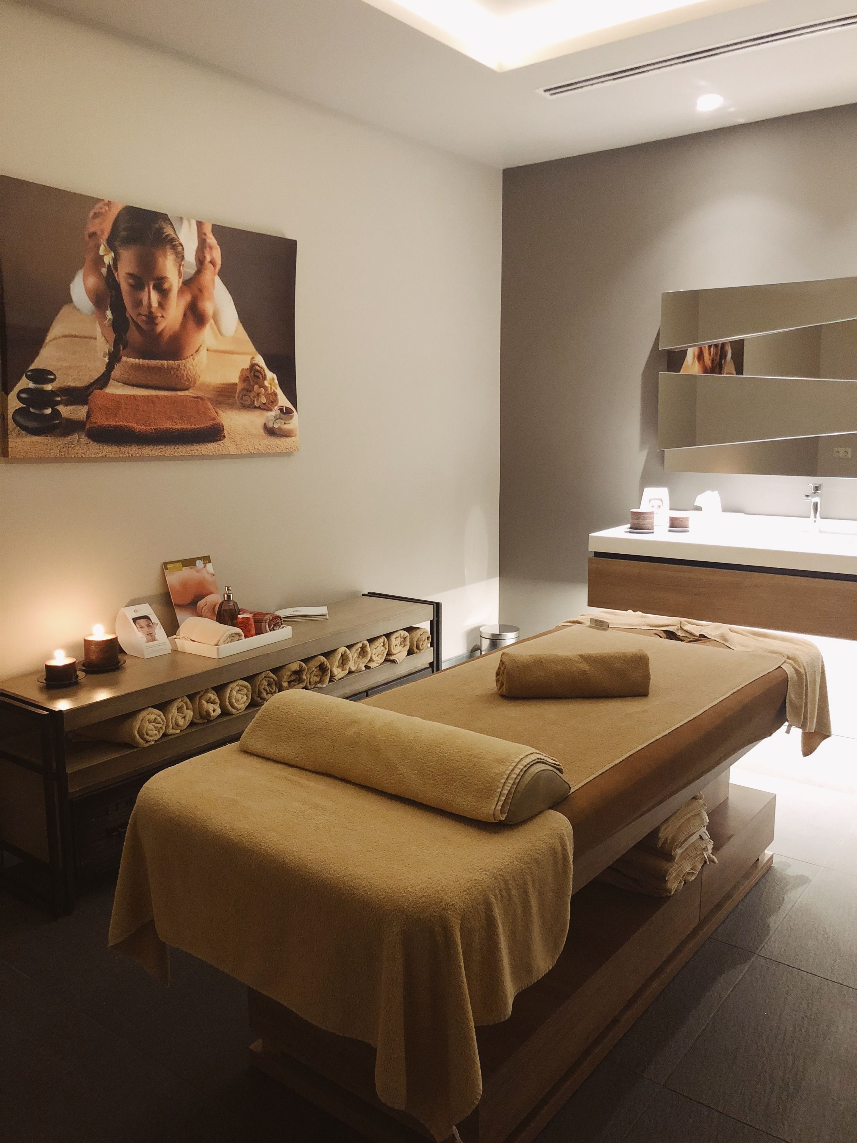 A spa room with a massage table, a wooden bench, a shelf with towels, a mirror, and a wall-mounted picture.