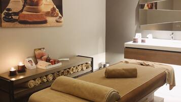 A spa room with a massage table, a wooden bench, a shelf with towels, a mirror, and a wall-mounted picture.