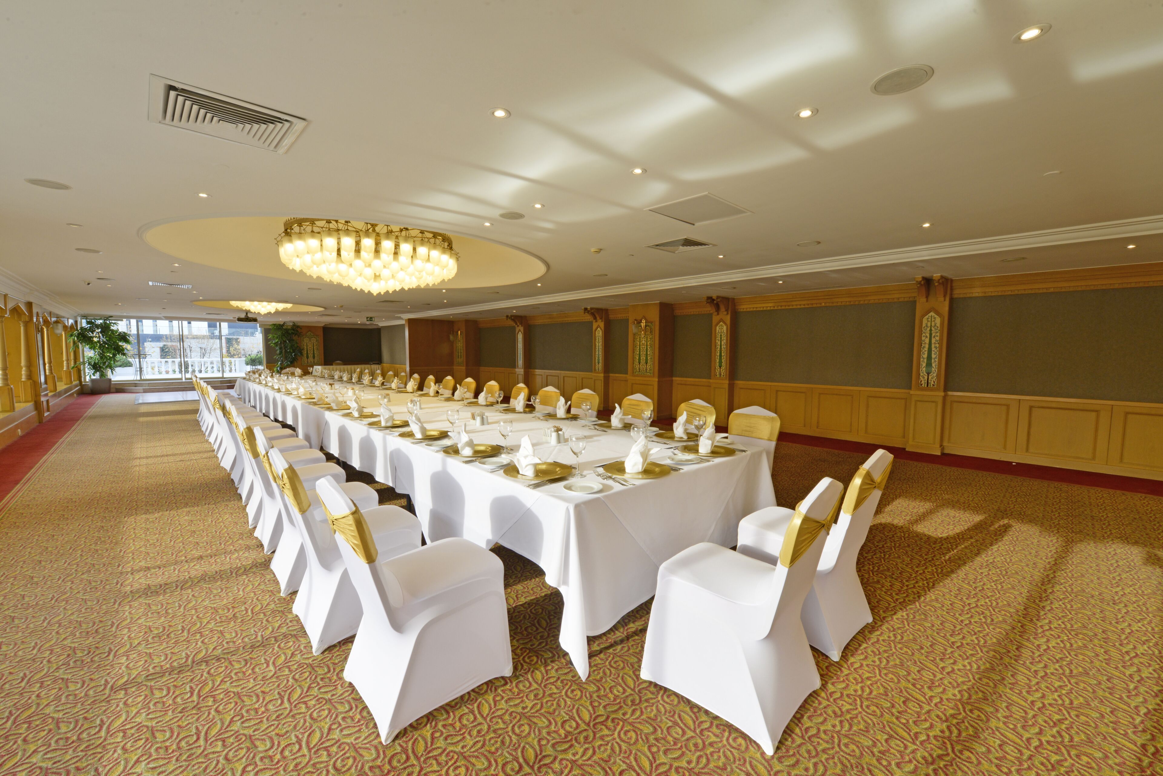 Un salón de banquetes con largas mesas dispuestas para un evento formal, con sillas blancas adornadas con cintas doradas, una alfombra estampada y una gran lámpara de araña.