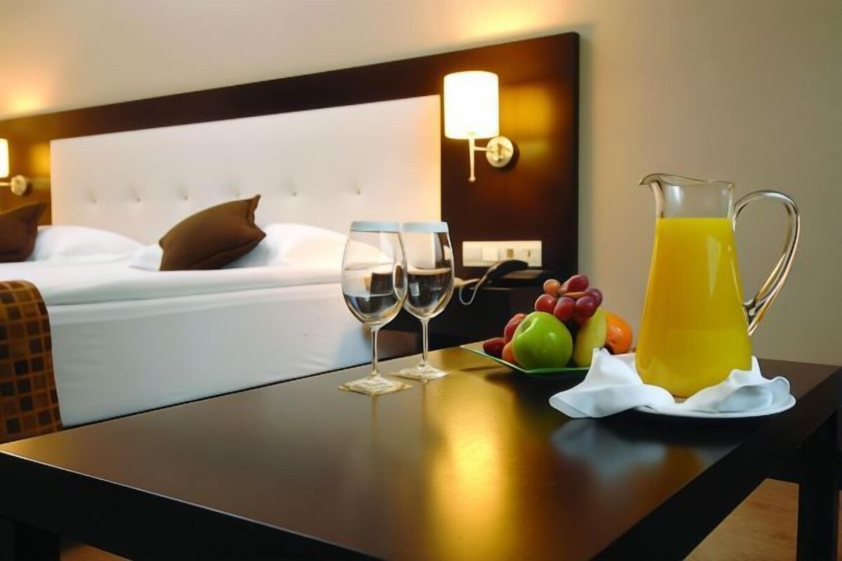 A hotel room with a neatly made bed, two wine glasses, a fruit bowl, and a pitcher of juice on a table.