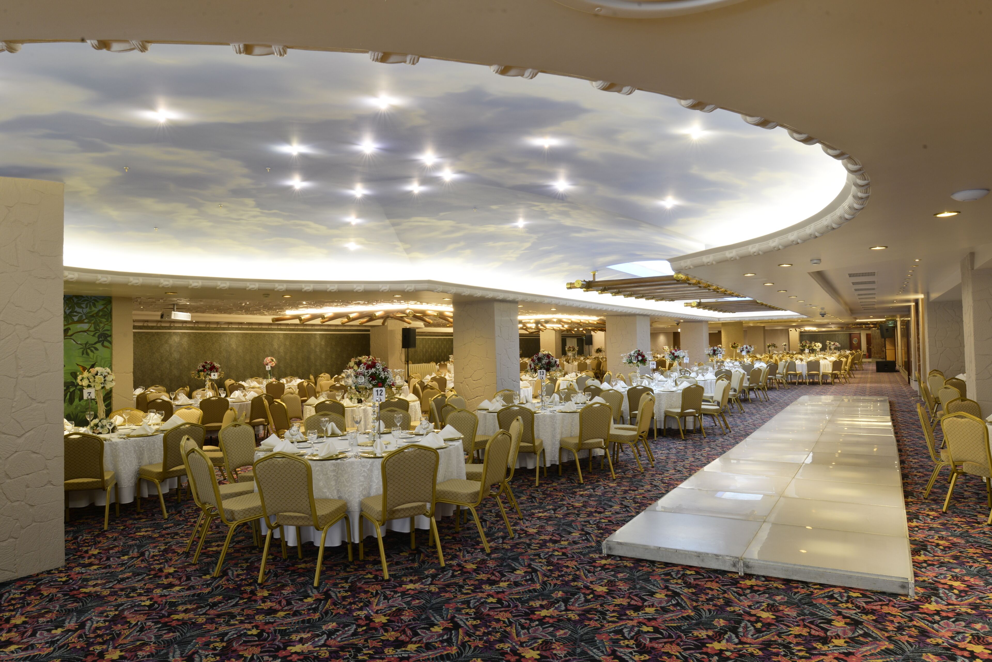 Un salón de banquetes con mesas redondas, sillas doradas y un techo decorado con un diseño de nubes.