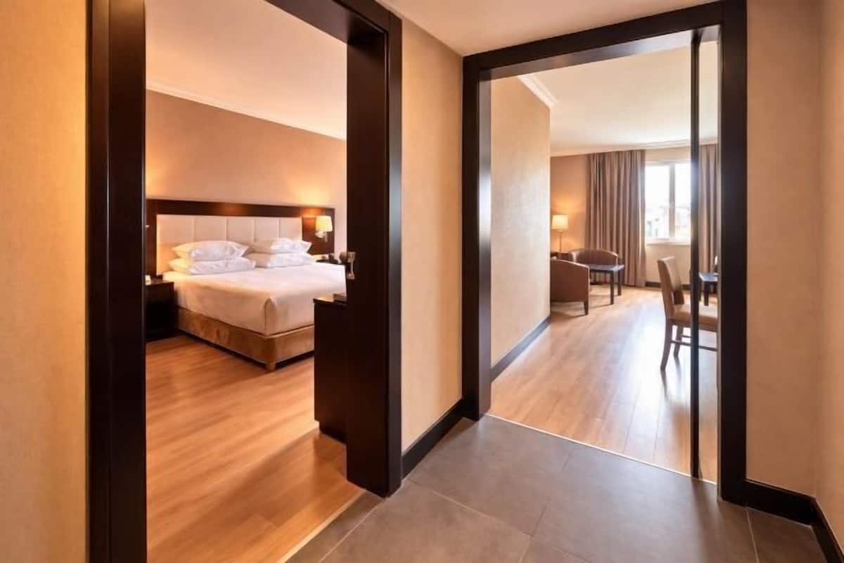 A hotel room with a bed, bedside table, and a view through a glass door to a dining area.