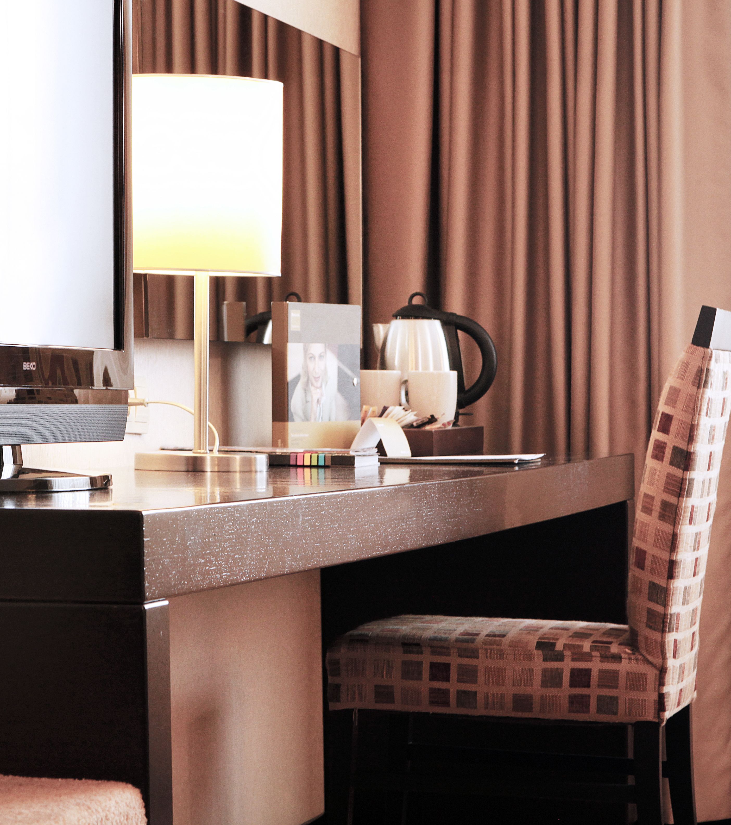 A hotel room with a desk, a lamp, a chair, a coffee maker, and a television.