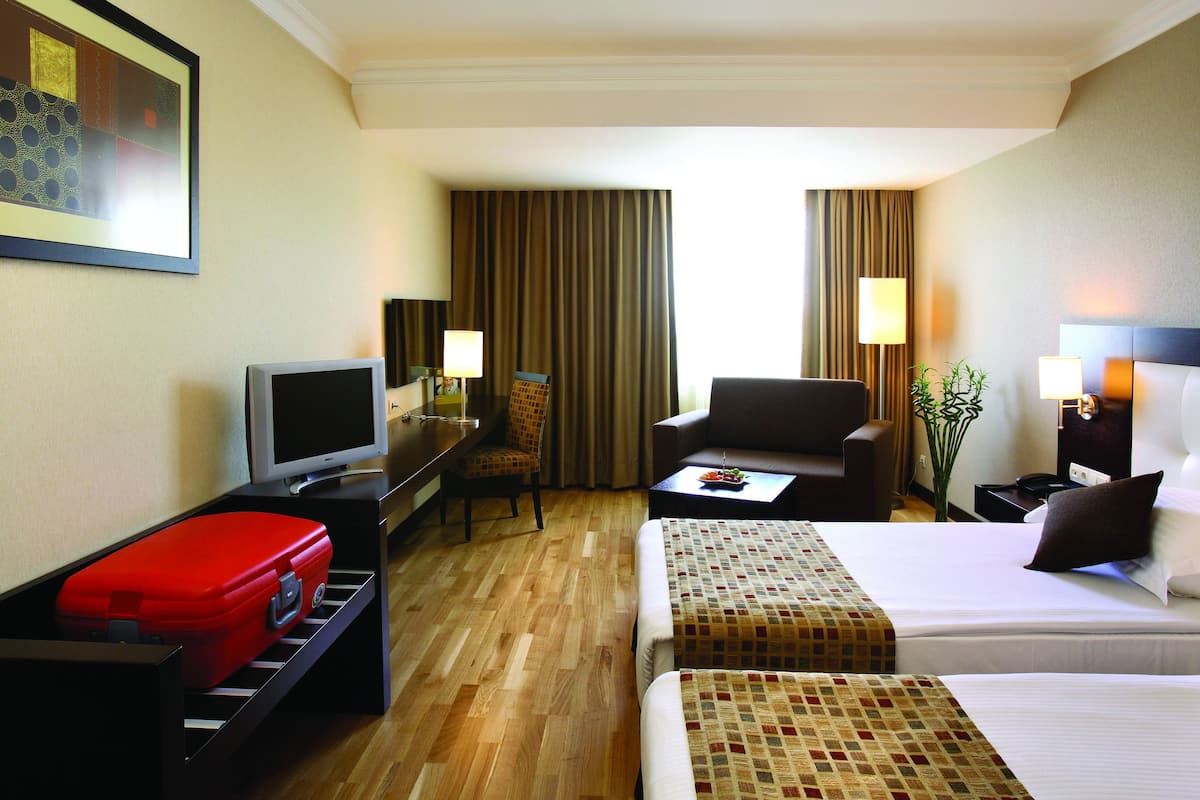 A hotel room with a bed, a desk with a computer, a chair, a red suitcase, and a television.