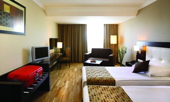 Habitación de hotel con una cama, un escritorio con computadora, una silla, una maleta roja y un televisor.