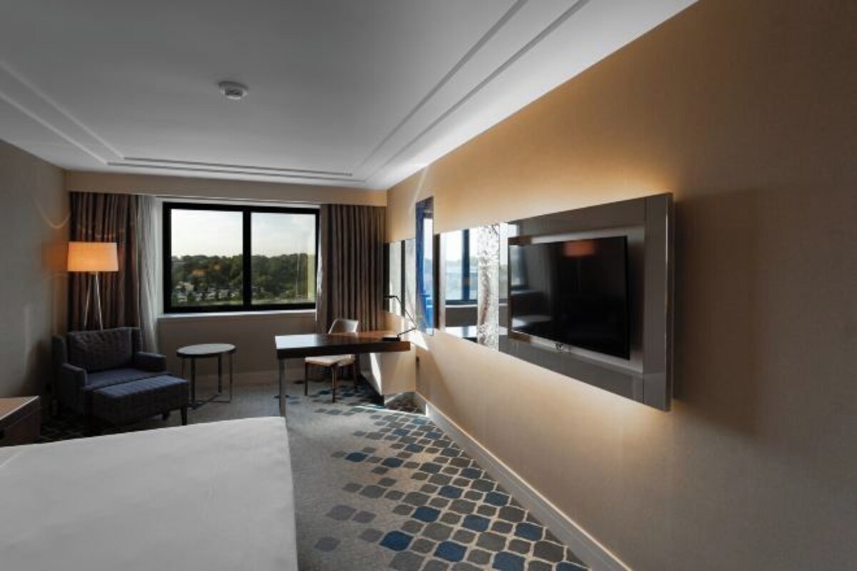 A hotel room with a bed, a desk, a chair, a TV mounted on the wall, and a window with a view.