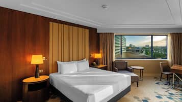 A hotel room with a large bed, two bedside lamps, a desk with a chair, and a view of the city through the window.