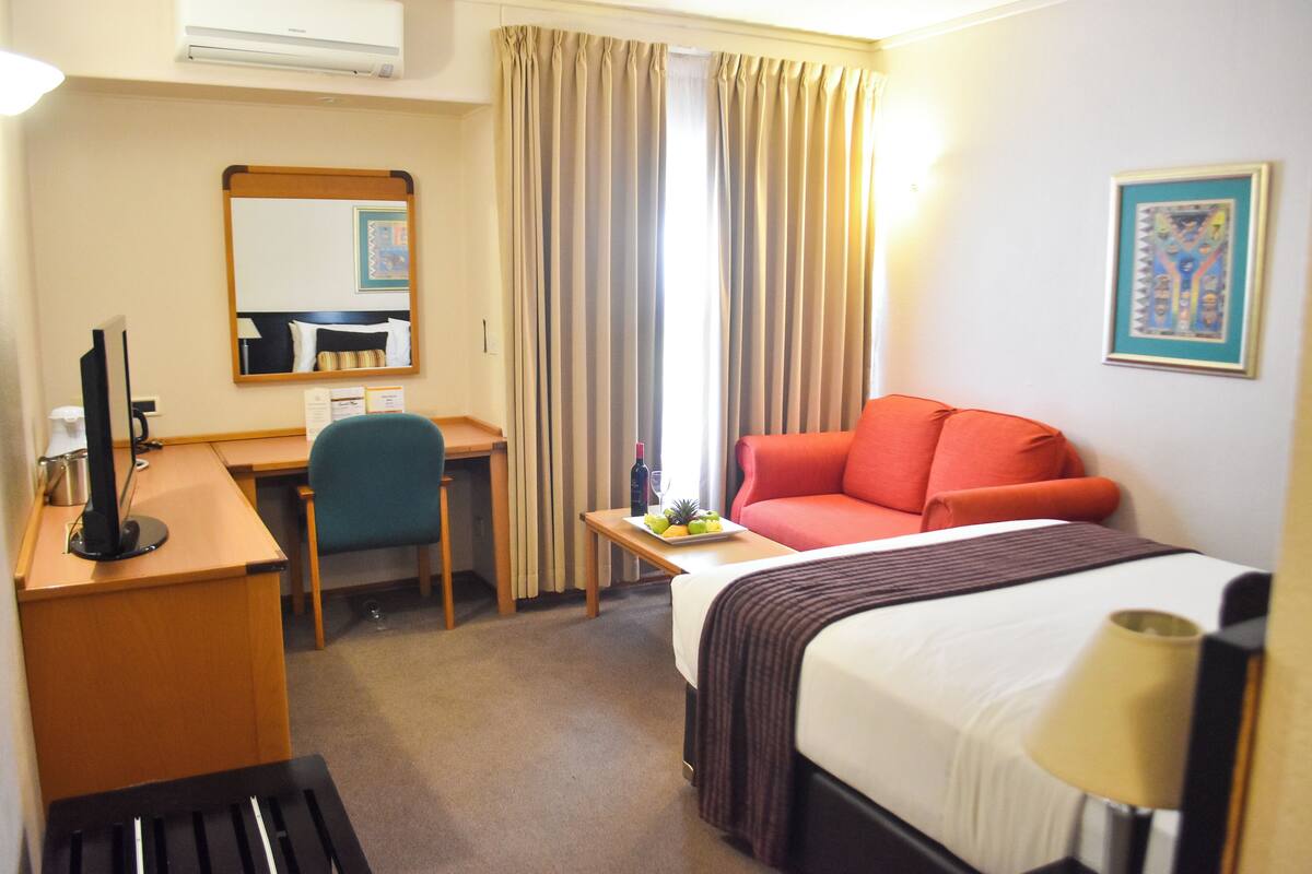 A hotel room with a bed, desk, chair, and a small table with fruit.