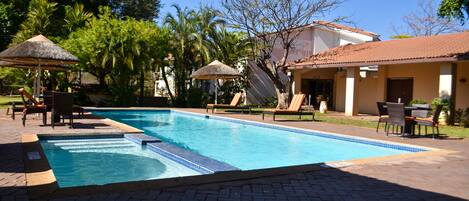 A swimming pool with lounge chairs and umbrellas, surrounded by palm trees and a building with a tiled roof.